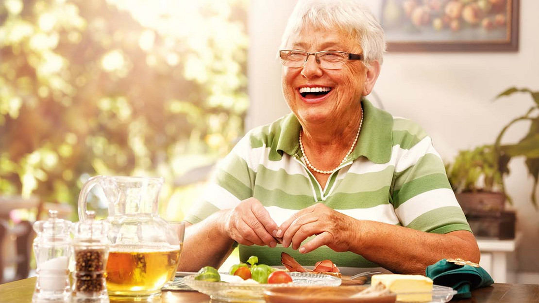 Ältere Frau sitzt lachend am gedeckten Frühstückstisch