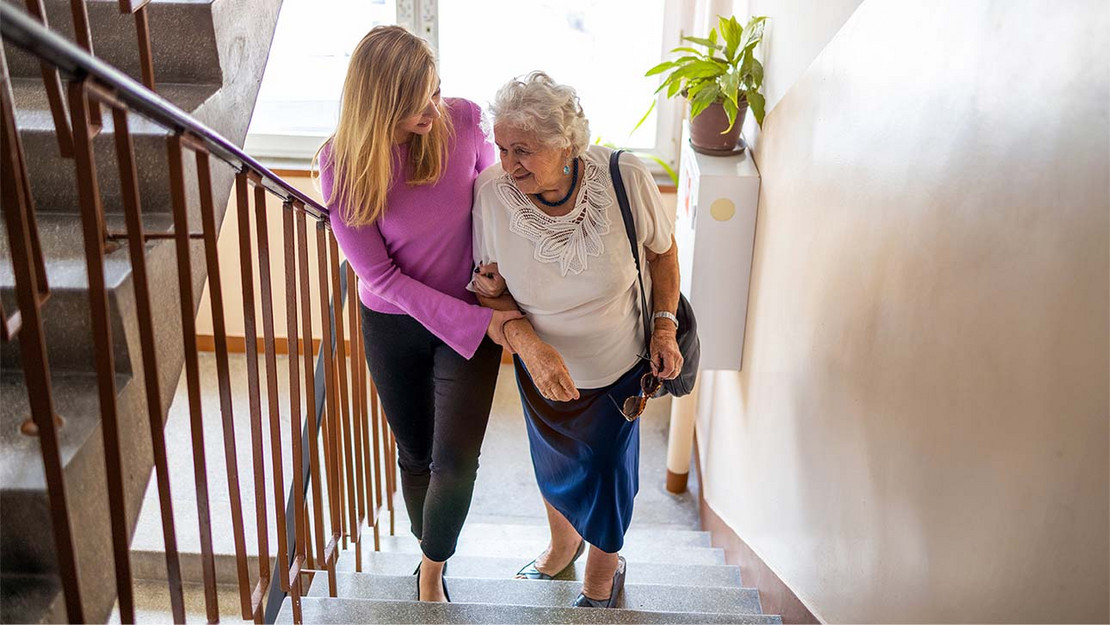 Eine jüngere Frau hält den Arm einer älteren Frau und geht gemeinsam mit ihr die Treppe in einem Wohnhaus herauf. 