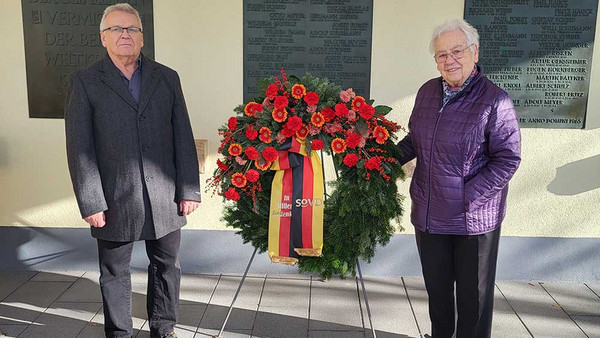 Mann und Frau auf Friedhof mit großem Kranz in der Mitte.