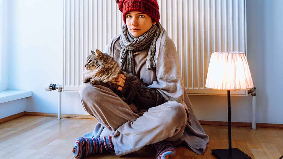 Frau mit Katze ist warm angezogen und sitzt vor einer Heizung. 