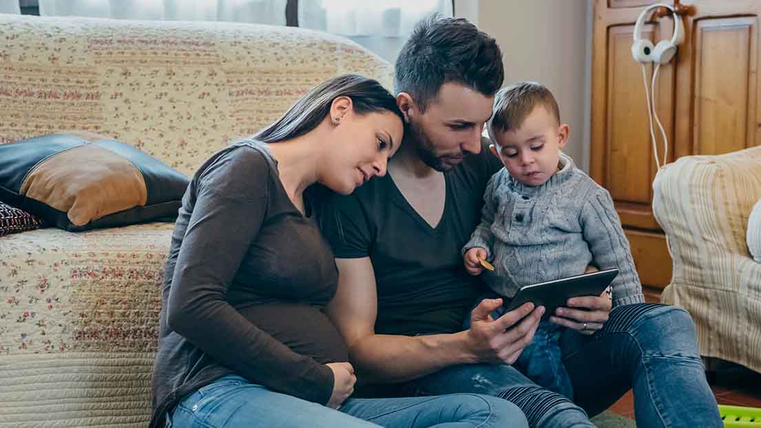 Familie sitzt vor dem Sofa auf dem Boden und schaut in ein Tablet. 