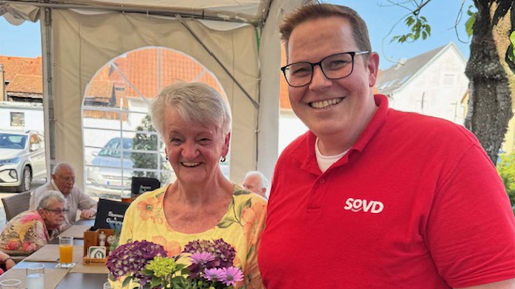 Ein Mann im SoVD-Shirt übergibt einer älteren Frau einen Blumenstrauß. 